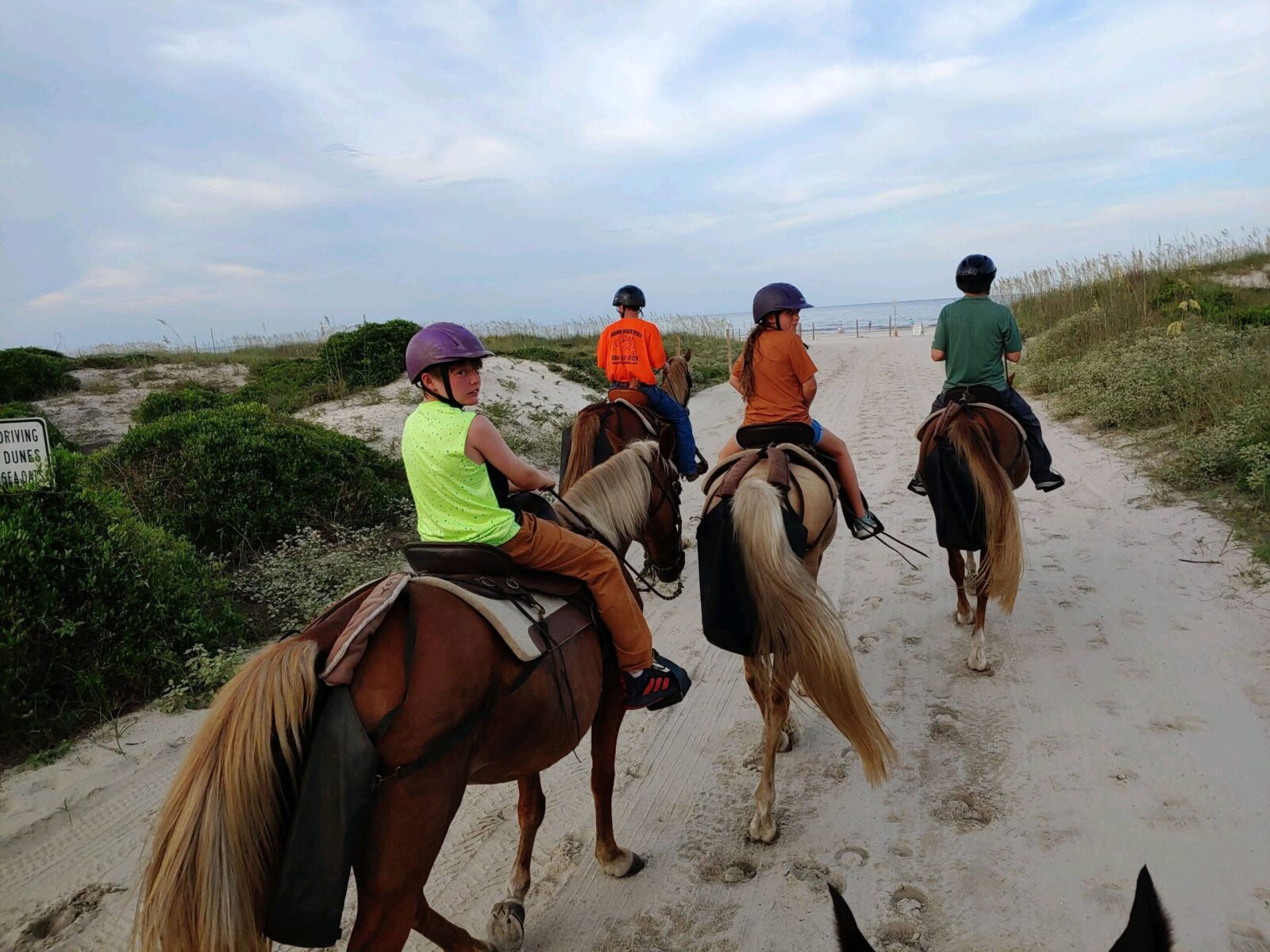 Four people horseback riding on a sandy path towards the beach, wearing helmets.