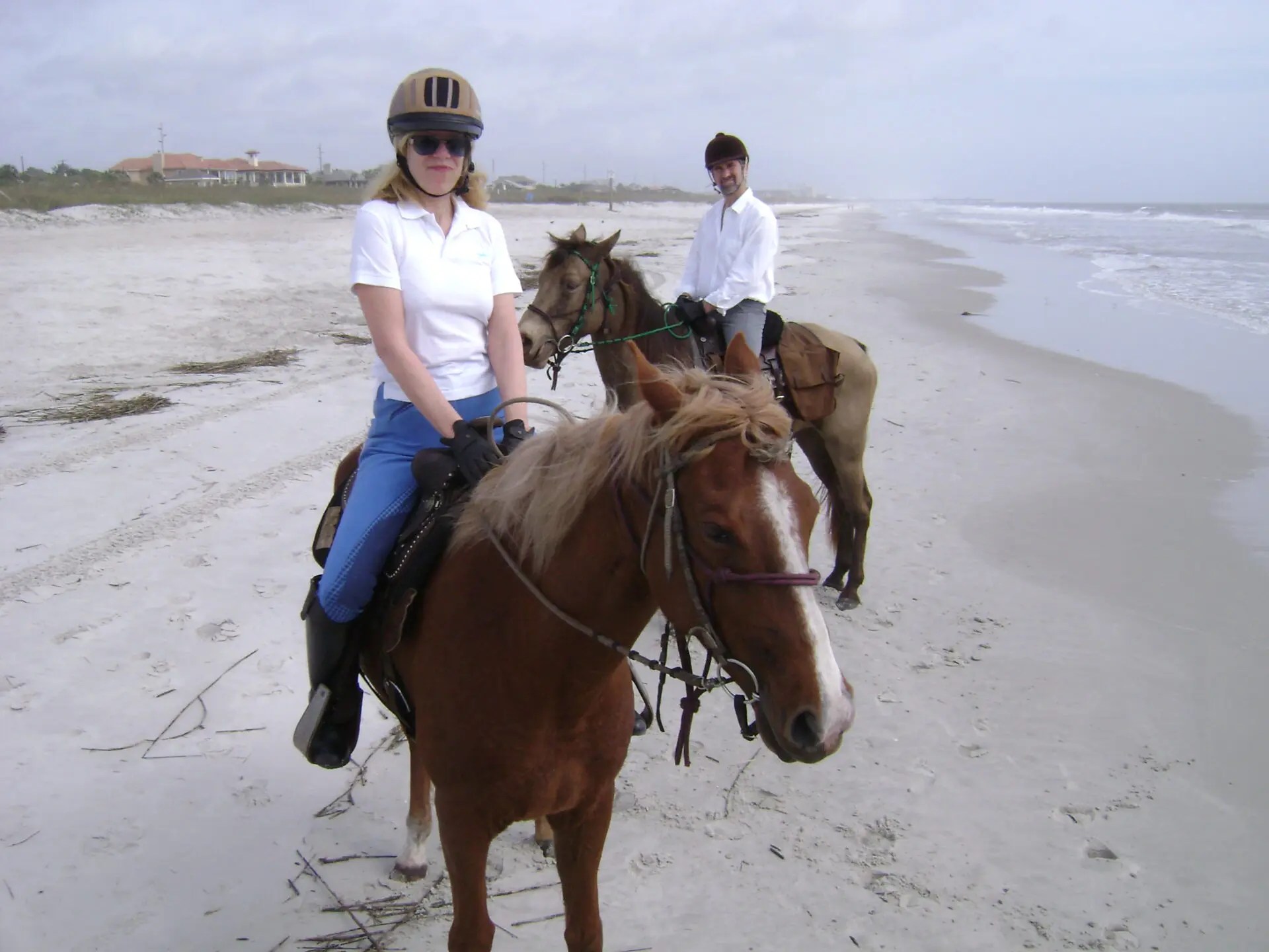 Two people riding horses on a sandy beach with ocean waves in the background.