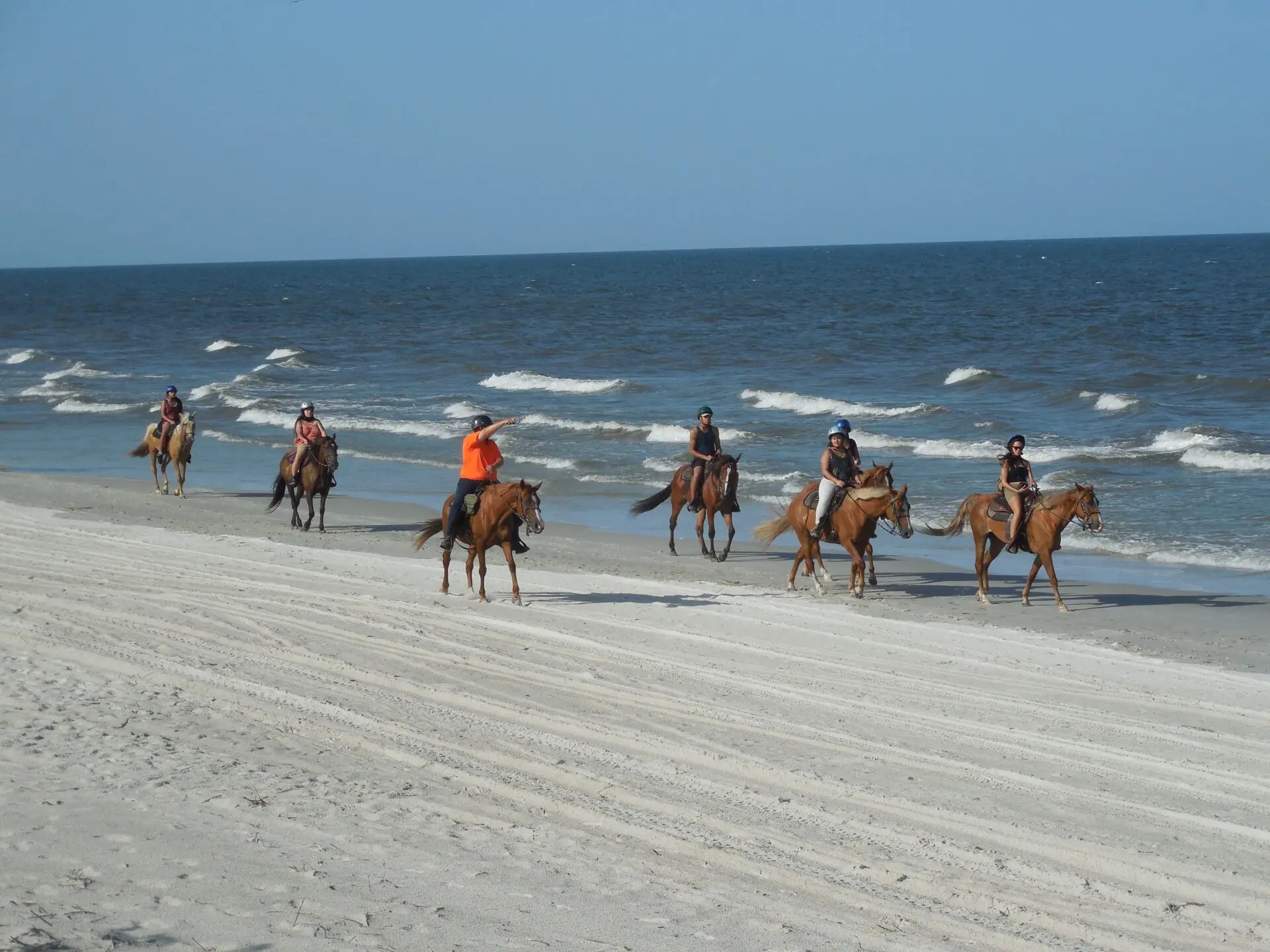 Group of people riding horses on a sandy beach by the ocean on a clear day.