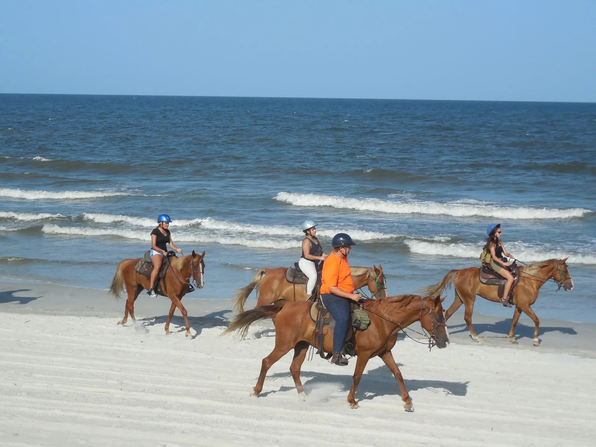 Four people riding horses on a sandy beach near the ocean on a sunny day.