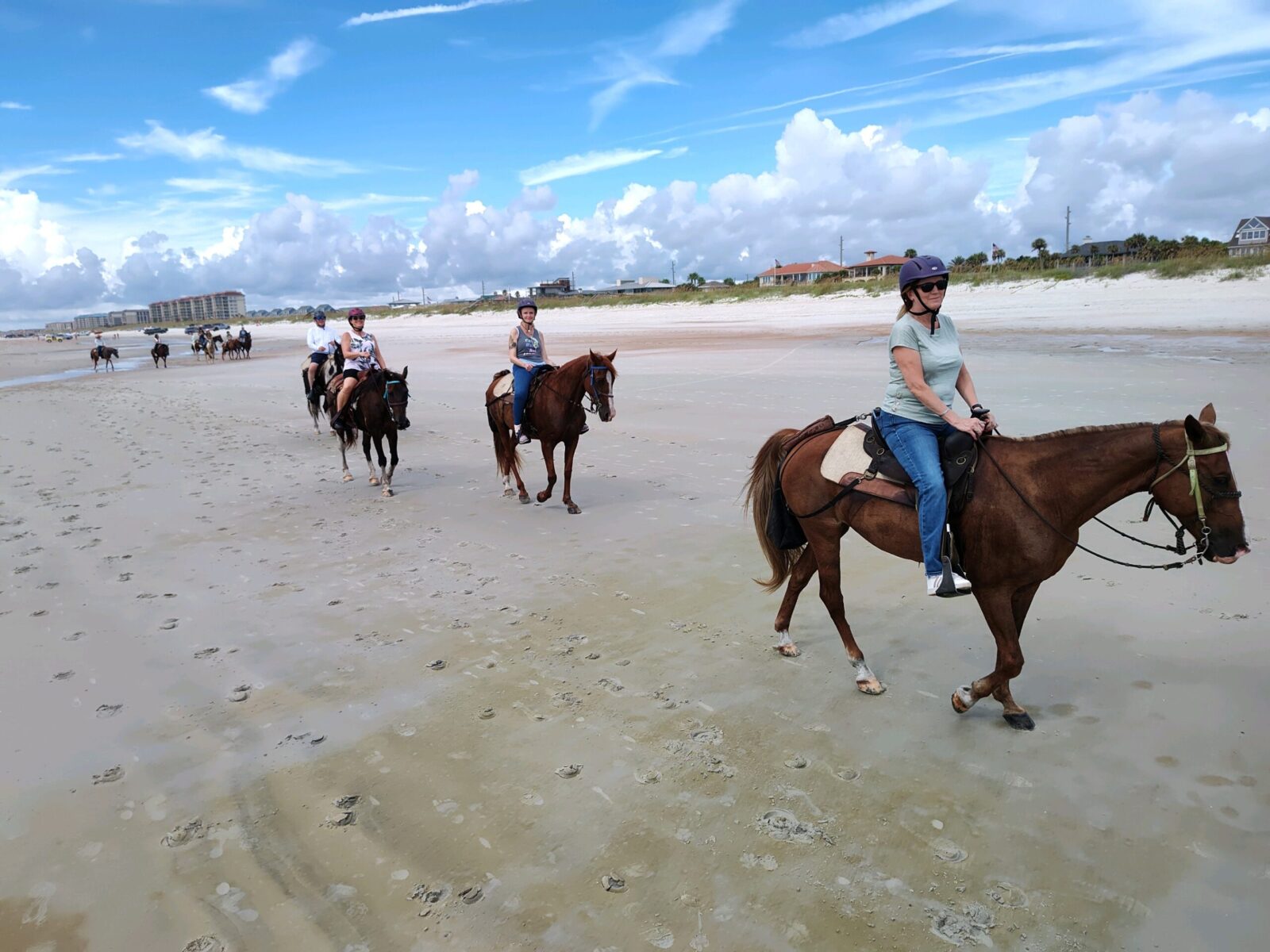 Group horseback riding on a sandy beach under a partly cloudy sky.