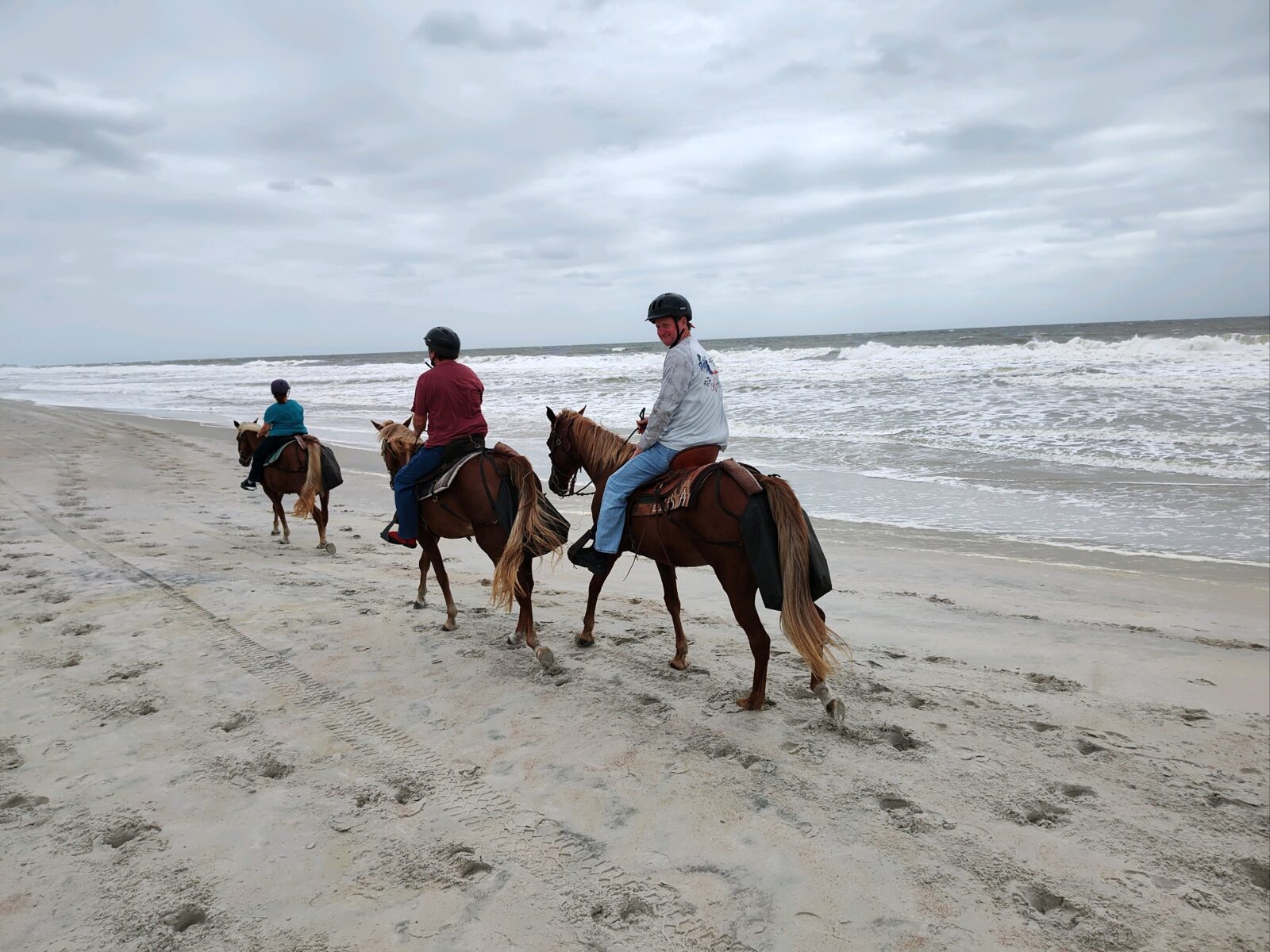 Three people horseback riding on a sandy beach by the ocean under cloudy skies.