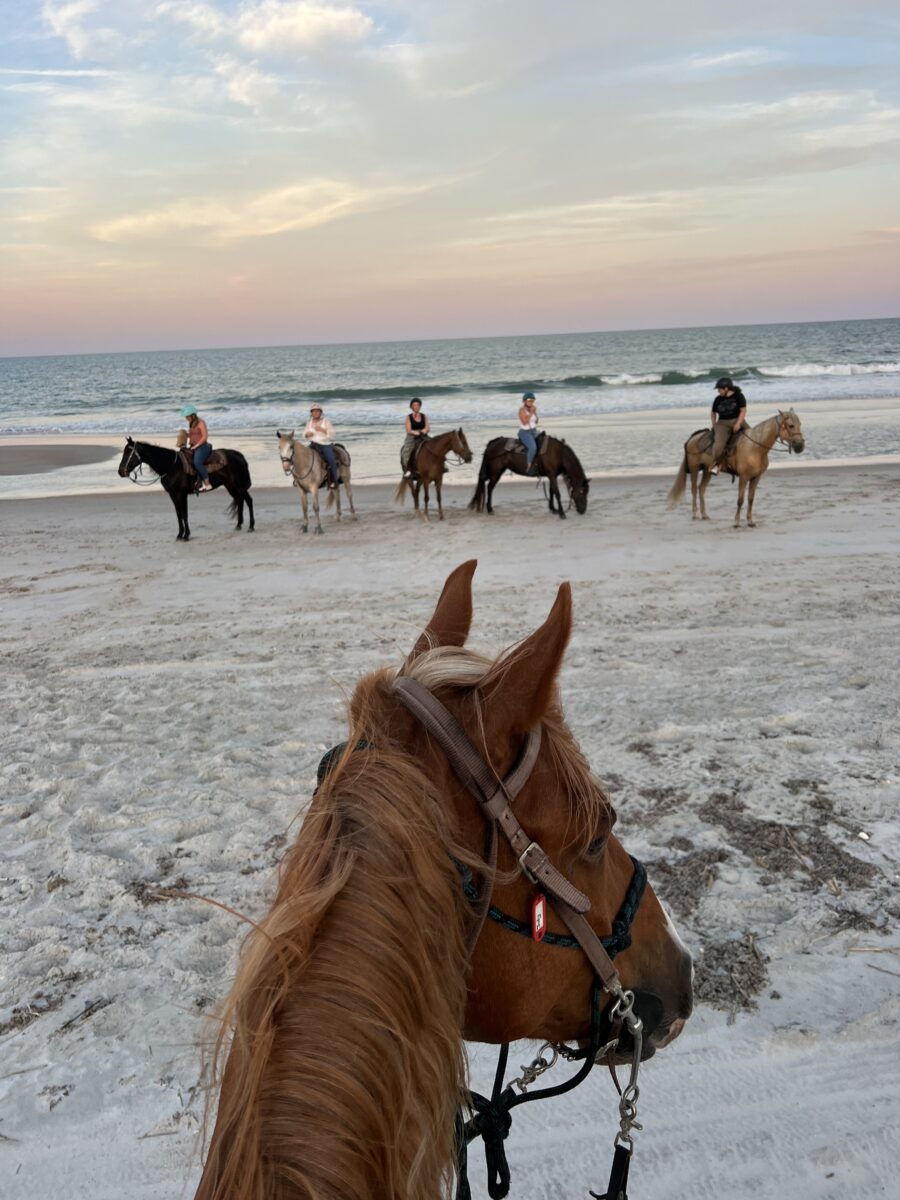 Horseback riders on sandy beach with view of ocean and sunset sky.