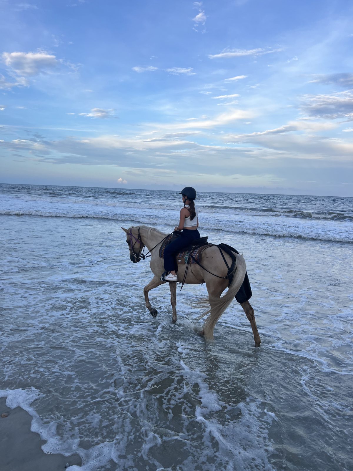 Person riding a horse along the beach with waves in the background and a blue sky above.
