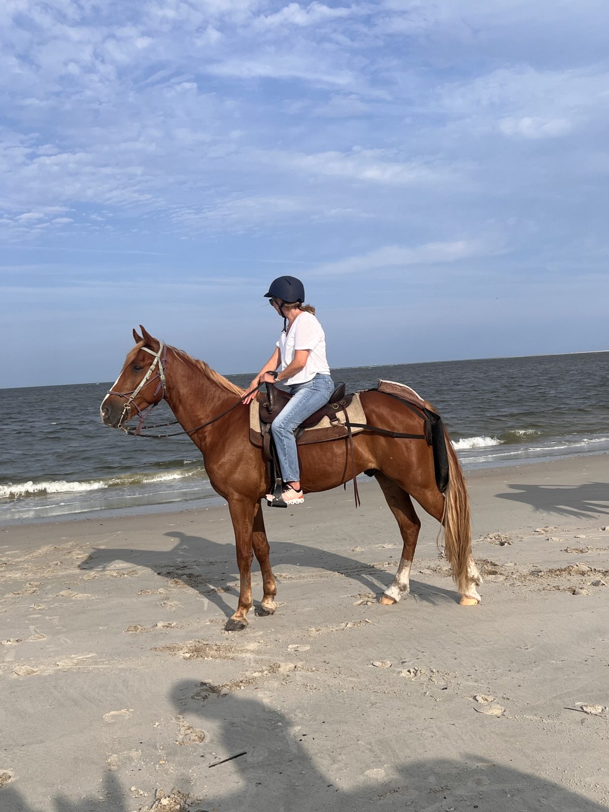 Person in helmet riding a horse on a sandy beach near the ocean.