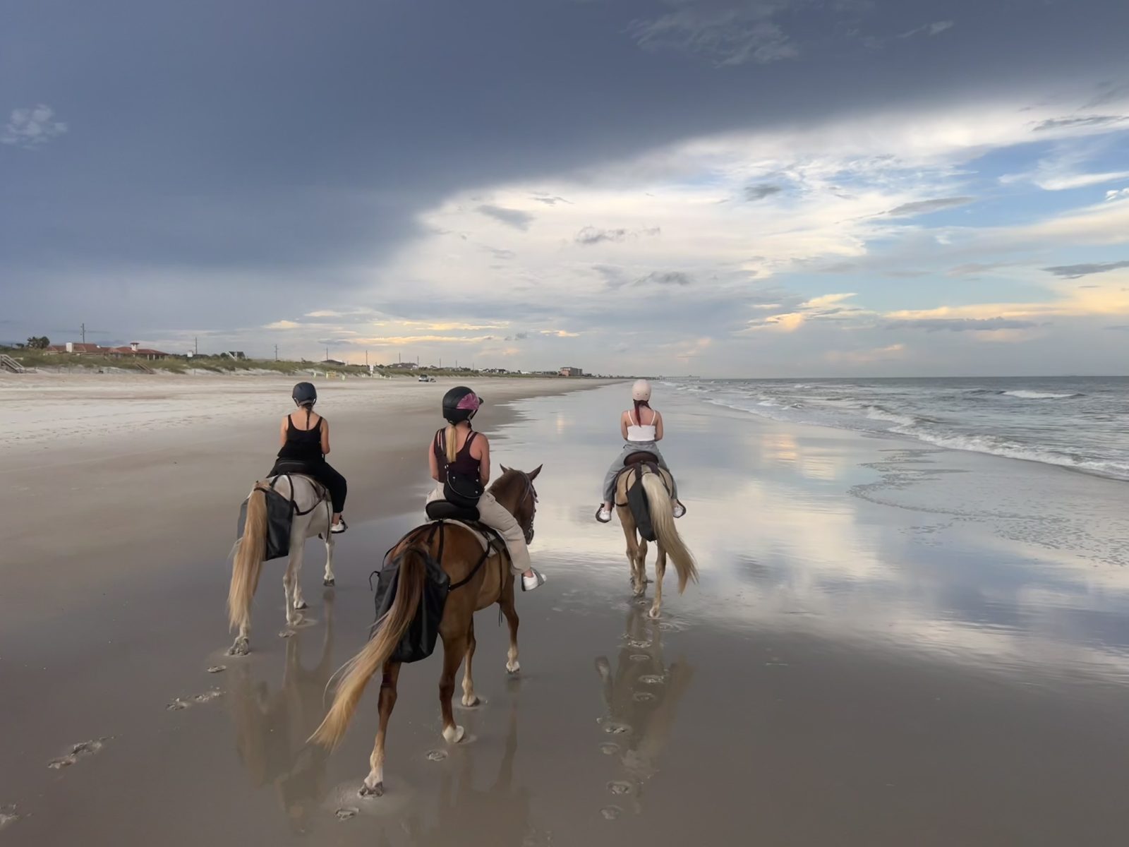 Three people riding horses on a beach with a cloudy sky and ocean waves.