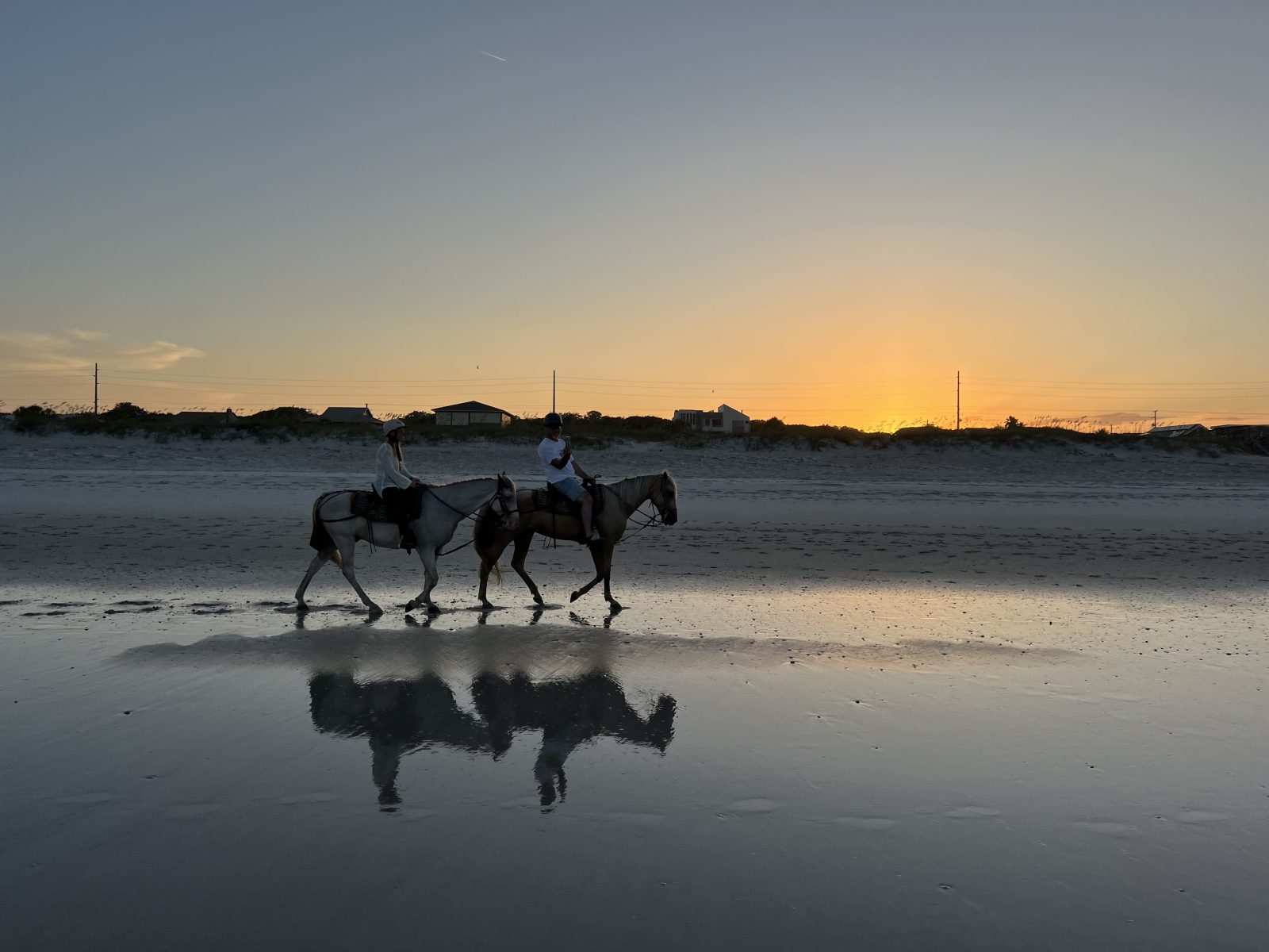 Two people horseback riding on a beach at sunset, with reflections on wet sand.