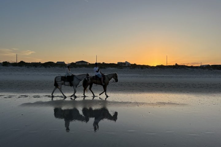 Two people horseback riding on a beach at sunset, with reflections on wet sand.