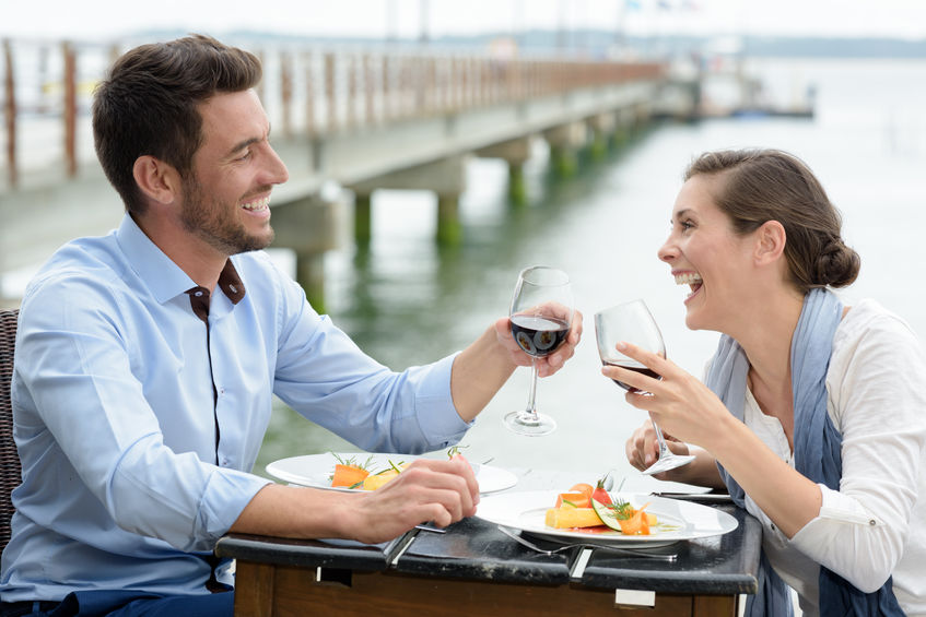A smiling couple clinking wine glasses while dining outdoors by the water.