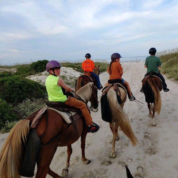 Four people riding horses on a sandy path near the beach, wearing helmets.