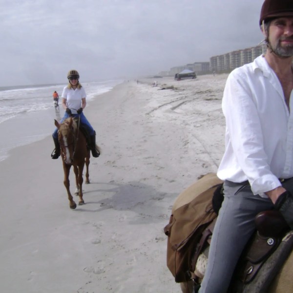 Two people horseback riding on a sandy beach near the ocean.