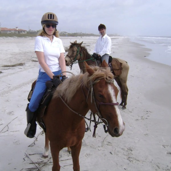 Two people riding horses on a sandy beach with ocean in the background.
