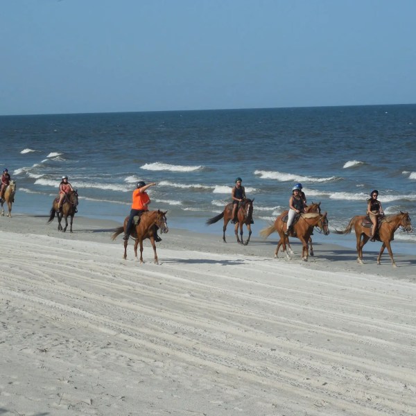 People riding horses along a beach with ocean waves in the background.
