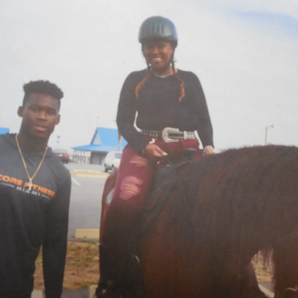 Person standing next to another on a horse, both smiling, with a building in the background.