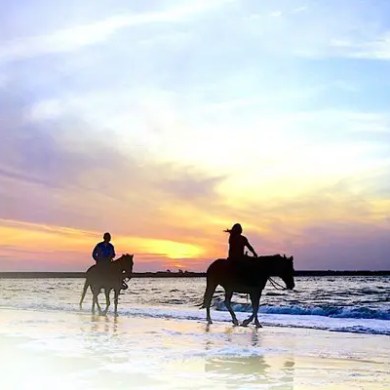 Two people horseback riding on a beach at sunset.