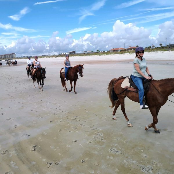 People horseback riding along a sandy beach under a blue sky with clouds.