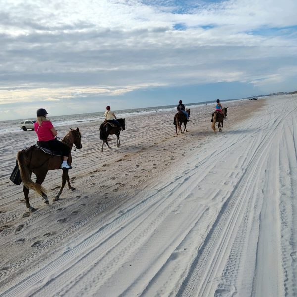 Four people riding horses along a sandy beach under a cloudy sky.