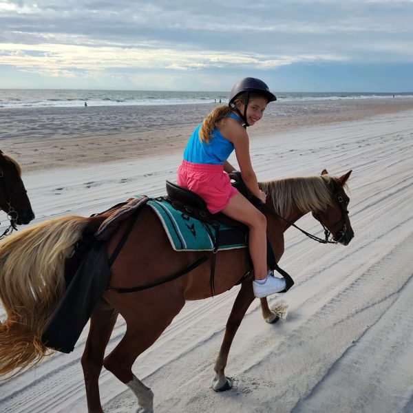 Girl riding a horse on a sunny beach, wearing a helmet and colorful clothes.