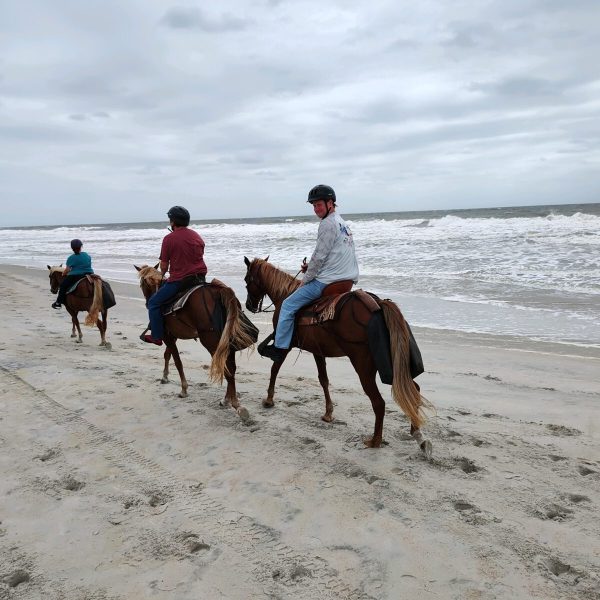 Three people riding horses along a sandy beach under a cloudy sky.