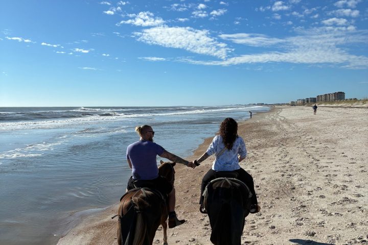 Two people horseback riding on a sunny beach, holding hands beside the ocean.