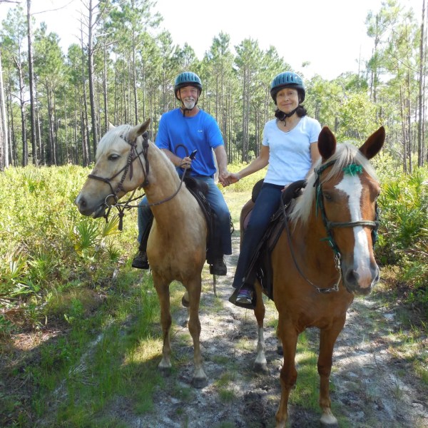 Two people on horses holding hands on a forest trail, wearing helmets, surrounded by trees and greenery.