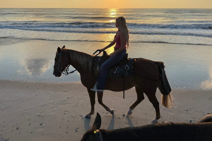 Person riding a horse on a beach at sunrise, with ocean waves and a cloudy sky.