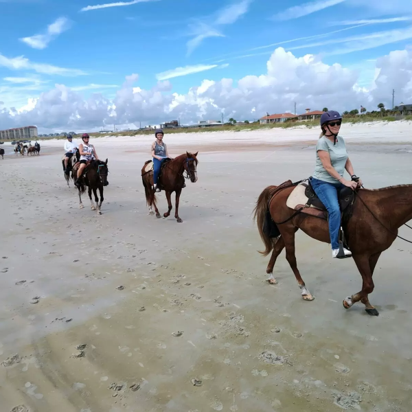 People riding horses on a sandy beach under a partly cloudy sky.