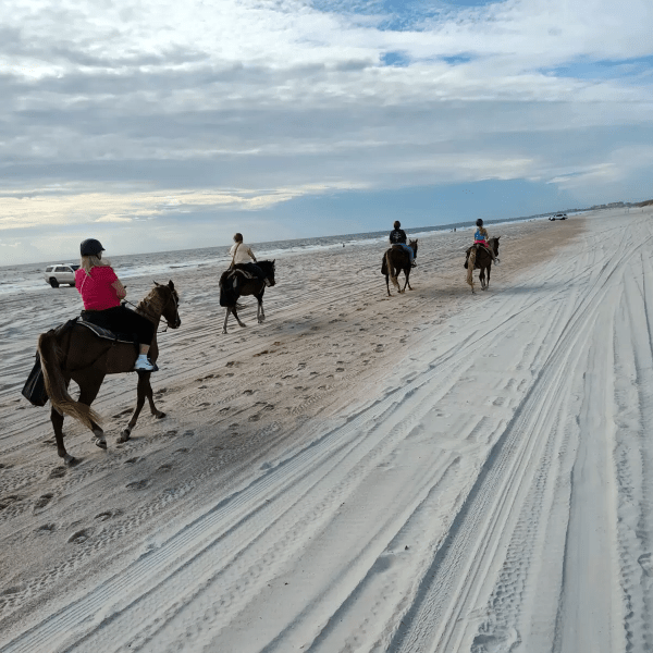 Four people riding horses on a sandy beach with a cloudy sky.